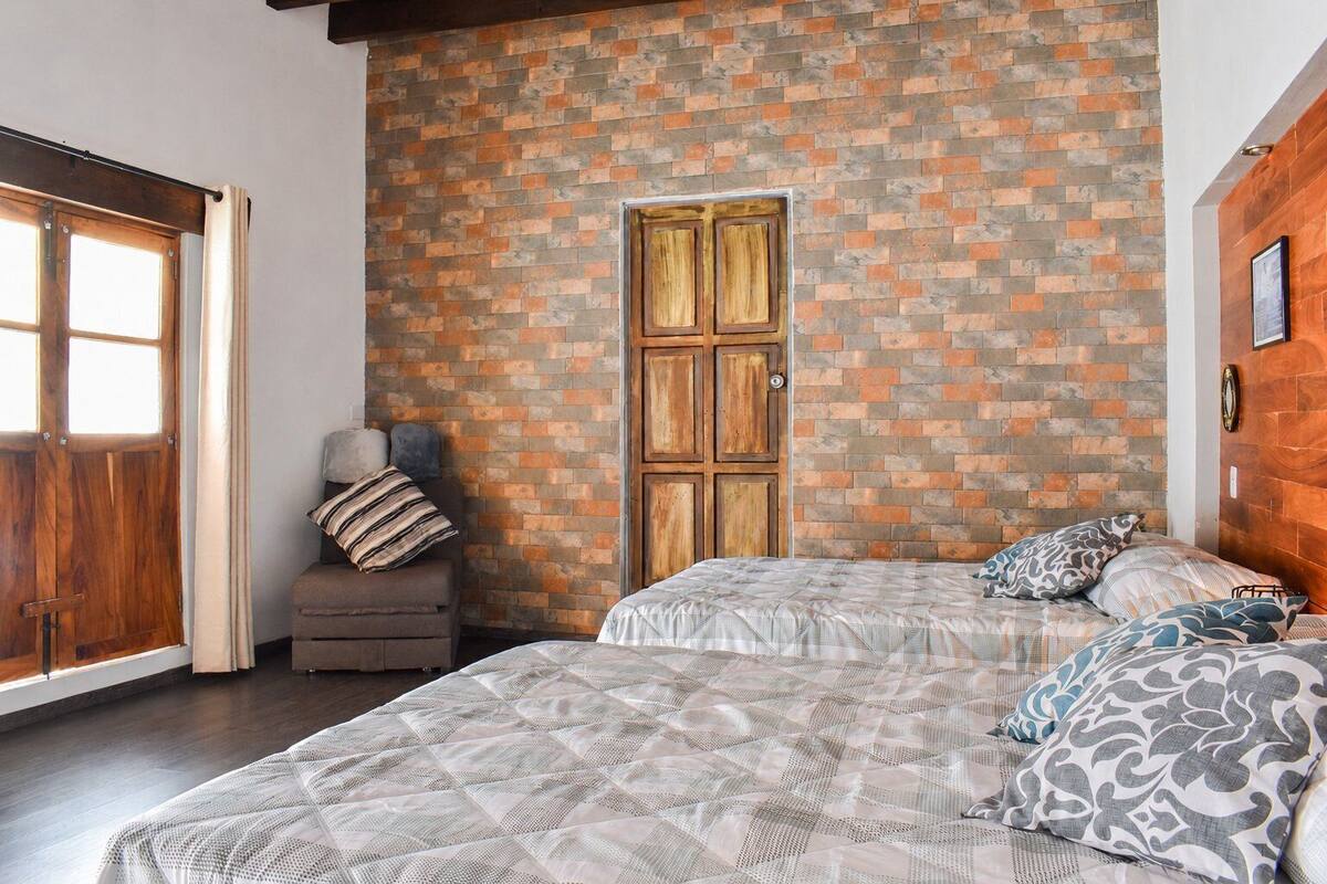 A bedroom with a brick-patterned wall, a wooden door, a sofa with cushions, and a bed with patterned bedding.
