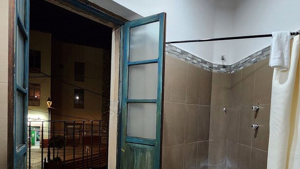 A room with a wooden floor, a door, a shower area, and a visible outdoor area with a street and buildings.