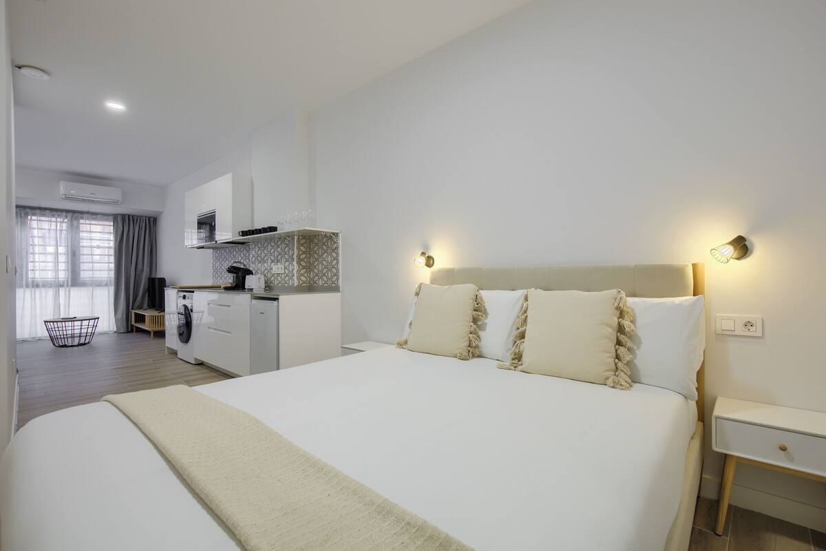 A modern hotel room with a large bed, a bedside table, a built-in washing machine, and a kitchenette.