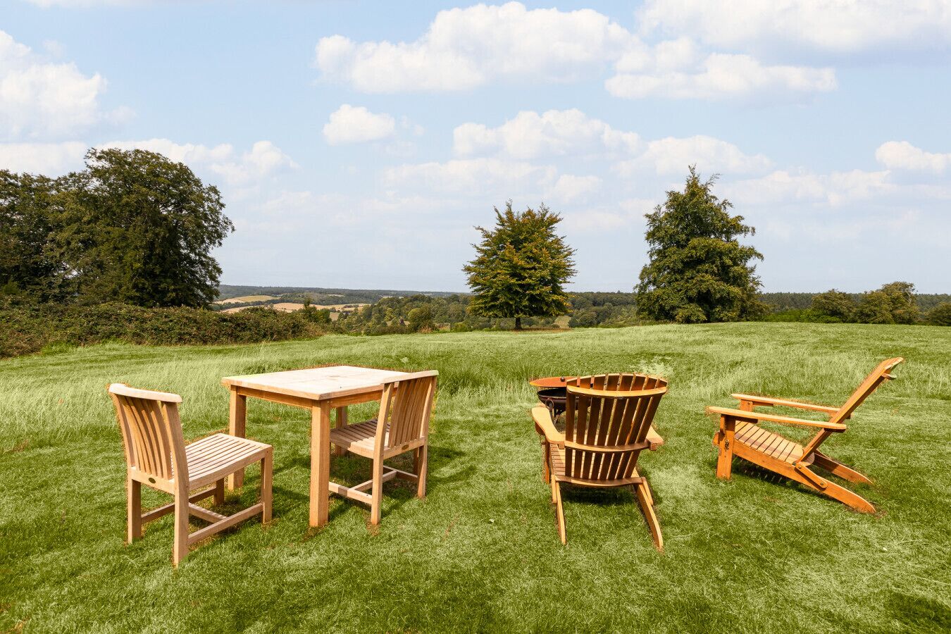 Ein Holztisch mit vier Stühlen auf einer Wiese, im Hintergrund Bäume und Hügel.