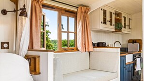 Un accogliente interno di una baita con una panca di legno, una finestra con tende, una piccola cucina con pensili e un letto.