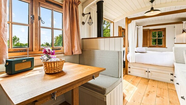 Una habitación acogedora con una mesa de madera, una radio y una cesta de flores. Hay una cama con dosel y una ventana con cortinas.