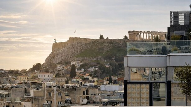 A rooftop terrace with wicker furniture, a glass balustrade, and a view of ancient ruins and modern buildings.