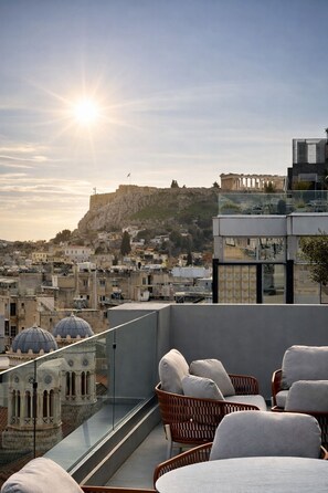 A rooftop terrace with wicker furniture, a glass balustrade, and a view of ancient ruins and modern buildings. - ACROPOLIS GRID ATHENS (Athens)