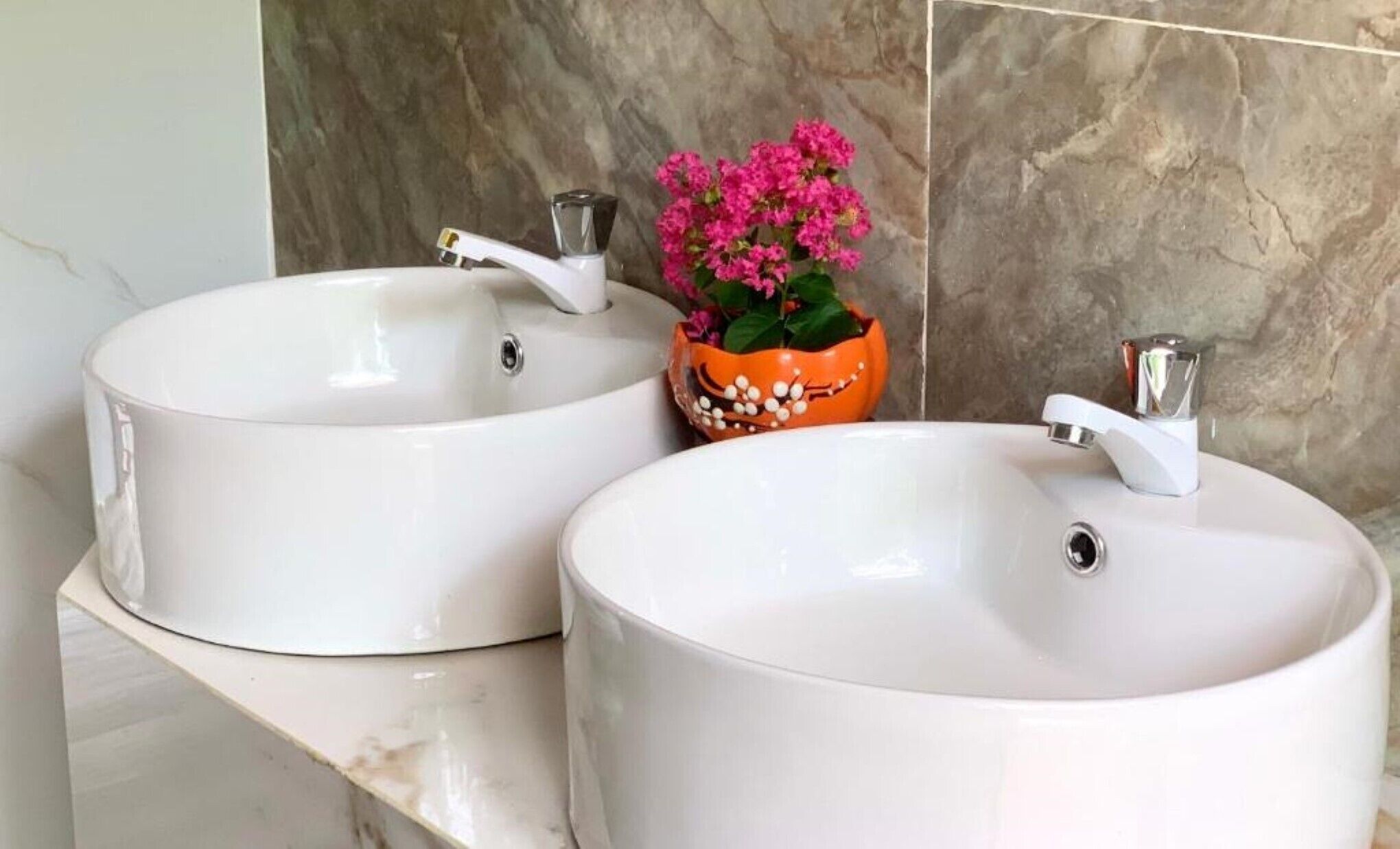 Two white sinks with chrome faucets, a marble countertop, and a decorative orange pot with pink flowers.