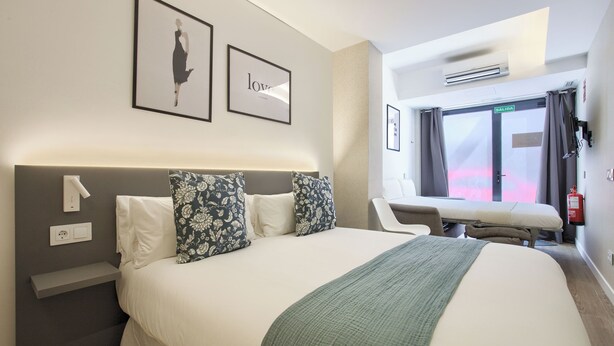 A hotel room with a large bed, a bedside table, a chair, a window with curtains, and a wall-mounted air conditioner. - Dobo Madrid Río (Madrid)