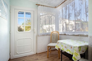 Una habitación con una puerta blanca, una ventana con cortinas de encaje, una silla de madera y una mesa con mantel floral.