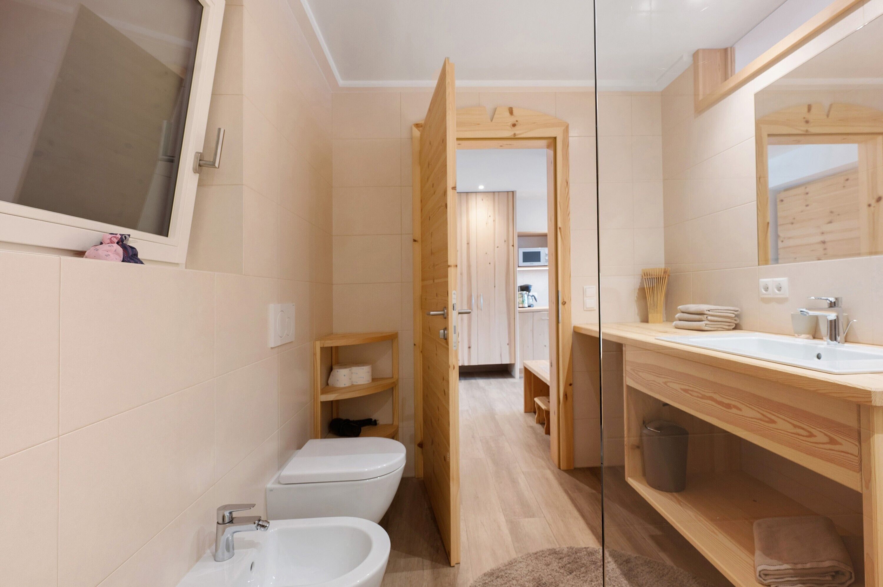 A modern bathroom with a toilet, bidet, and sink, wooden shelving, and a mirror.