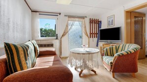 A furnished room with a sofa, armchair, TV, and a window with curtains. - Bungalow 'Manni' with Shared Terrace, Garden, and Wi-Fi (Koserow)