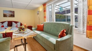 A cozy living room with a sofa, a table with a fruit bowl, a painting on the wall, and a window with curtains. - Bungalow 'Nati' with Shared Terrace, Garden, and Wi-Fi (Koserow)