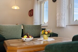 Een kamer met een houten tafel, een groene bank, een raam met gordijnen en een wandklok.