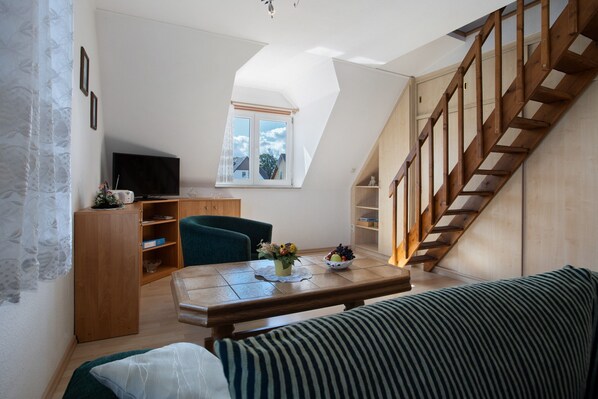 A loft-style room with a wooden staircase, a dining table with a vase, a sofa, and a television on a cabinet.