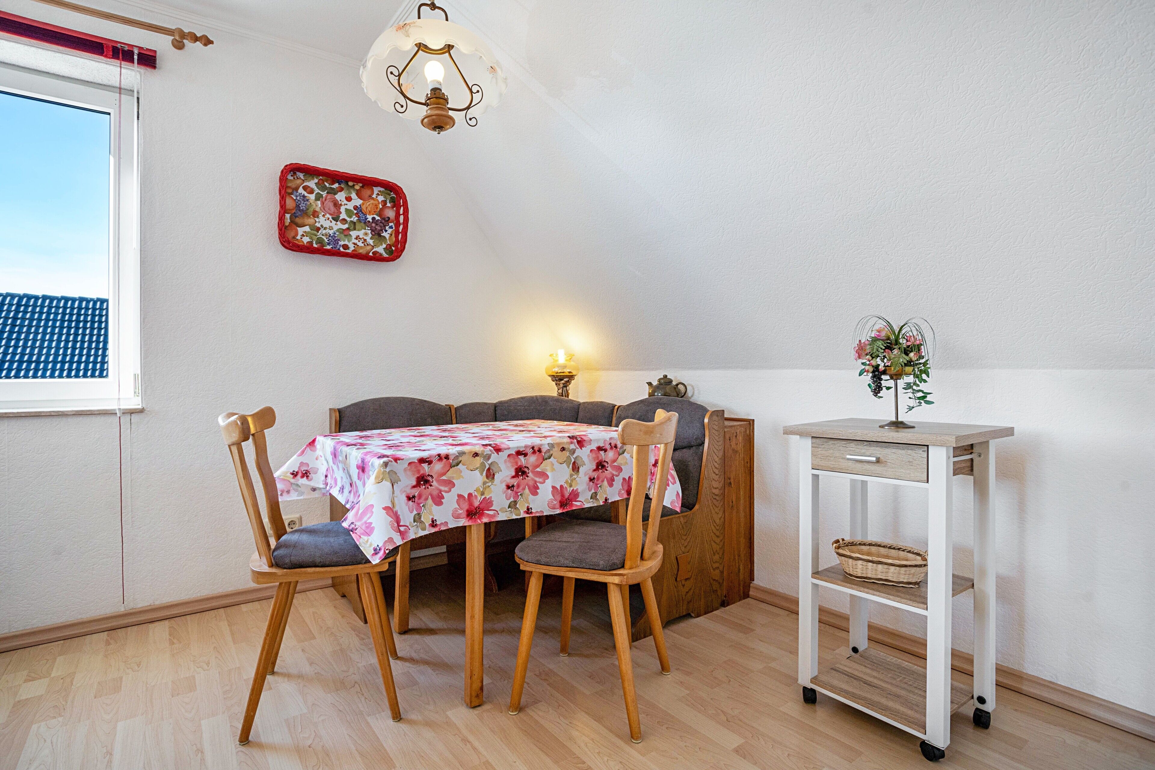 En matplats med ett bord, stolar, en lampa, en väggdekoration och en liten vit hylla med en växt.