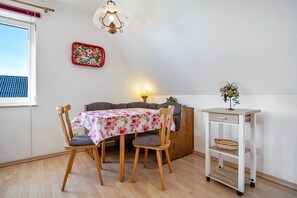 Uma área de refeições com mesa, cadeiras, um abajur, decoração na parede e uma pequena estante branca com uma planta.