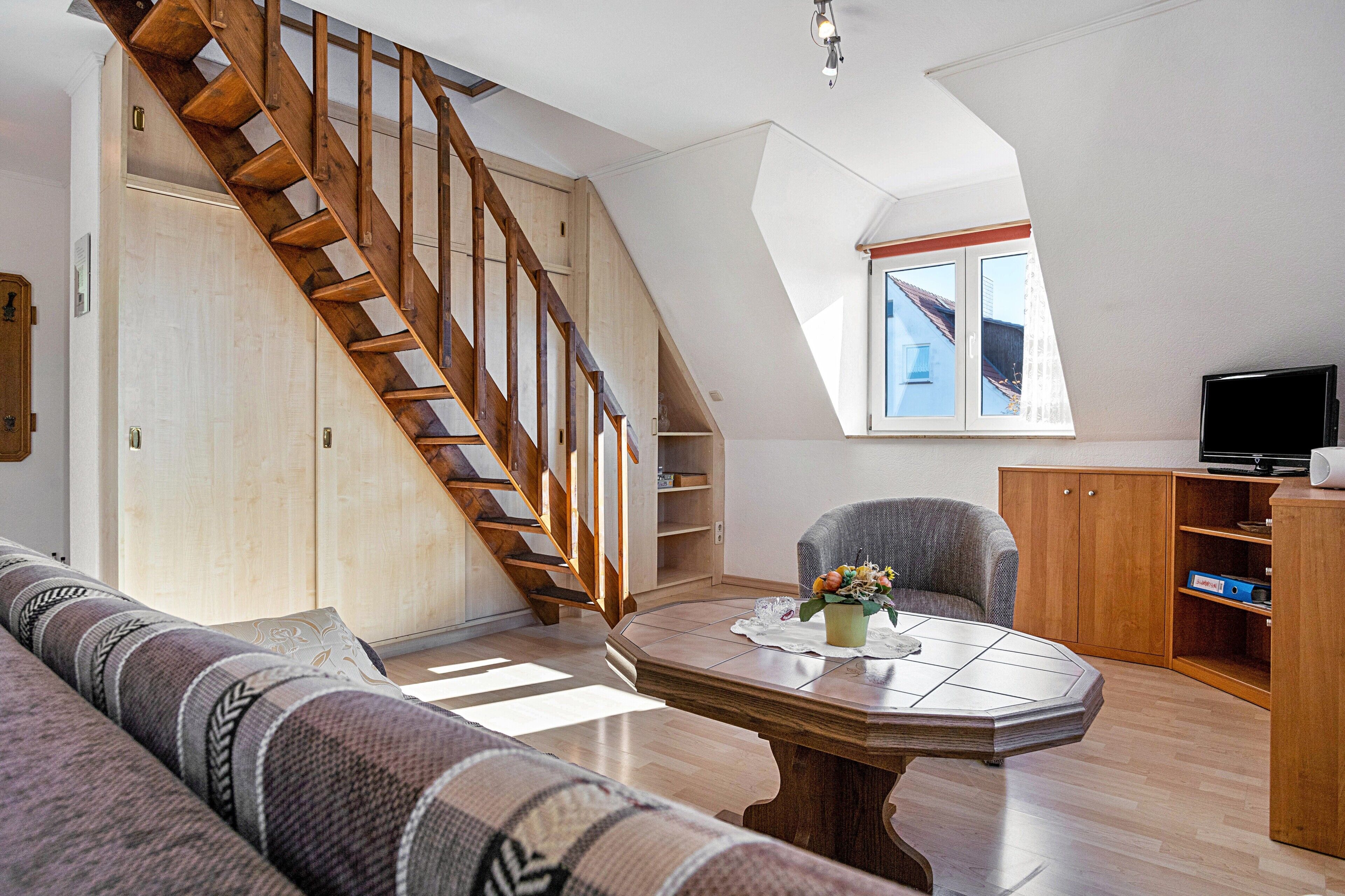 A spacious living room with a wooden staircase, a dining table, a sofa, and a television.