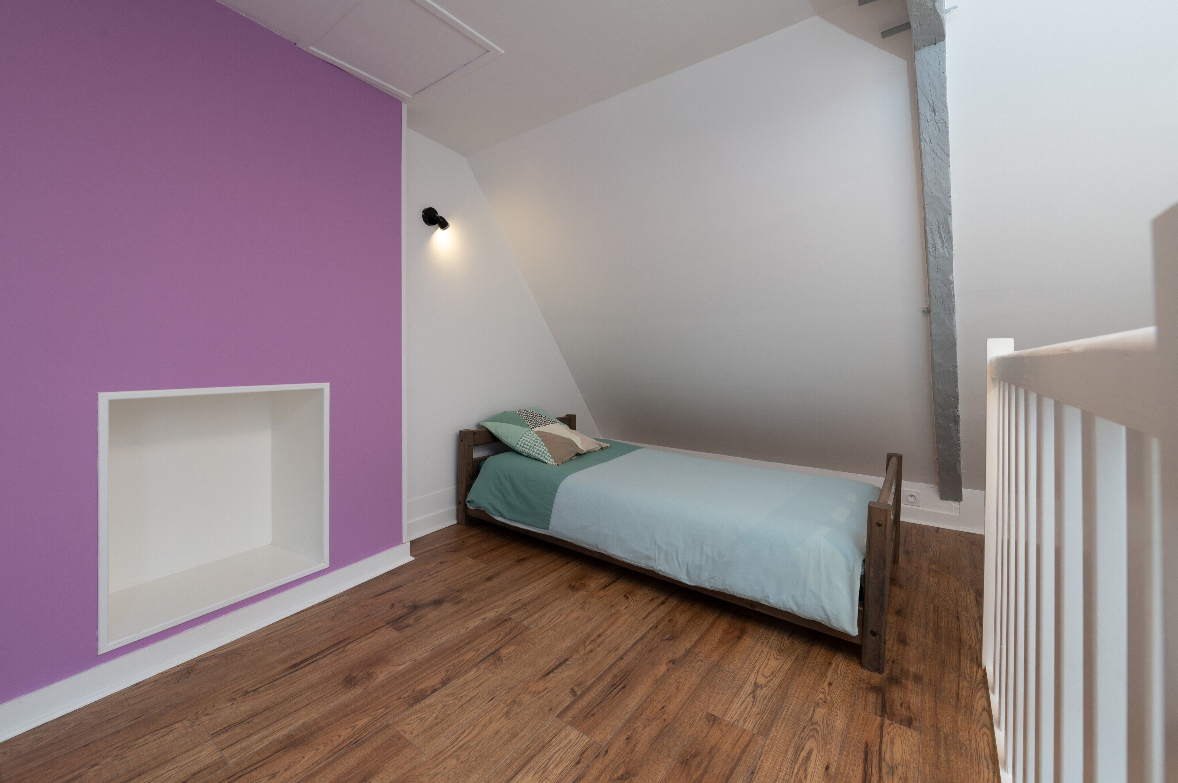 Una habitación con techo inclinado, una cama con estructura de madera, un estante blanco y una lámpara fijada a la pared.