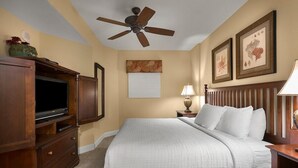Una habitación de hotel con una cama, un ventilador de techo, un televisor, un armario de madera y obras de arte enmarcadas en las paredes.