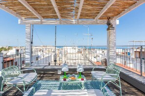 Une terrasse sur le toit agrémentée d’une pergola, de deux fauteuils de relaxation, d’une petite table avec un plateau en verre, et offrant une vue sur le paysage urbain et la mer.