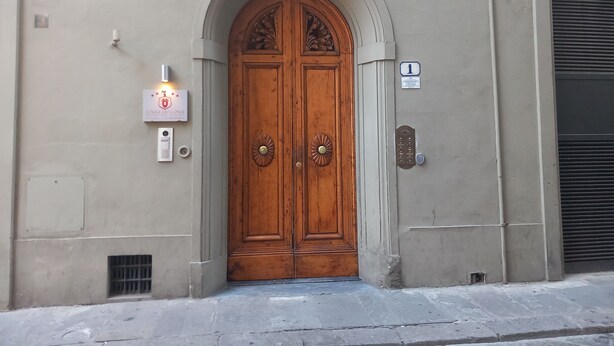 A double wooden door with a decorative arch above it, a wall-mounted light, and a doorbell. - Conti Apartment in Firenze (Florence)