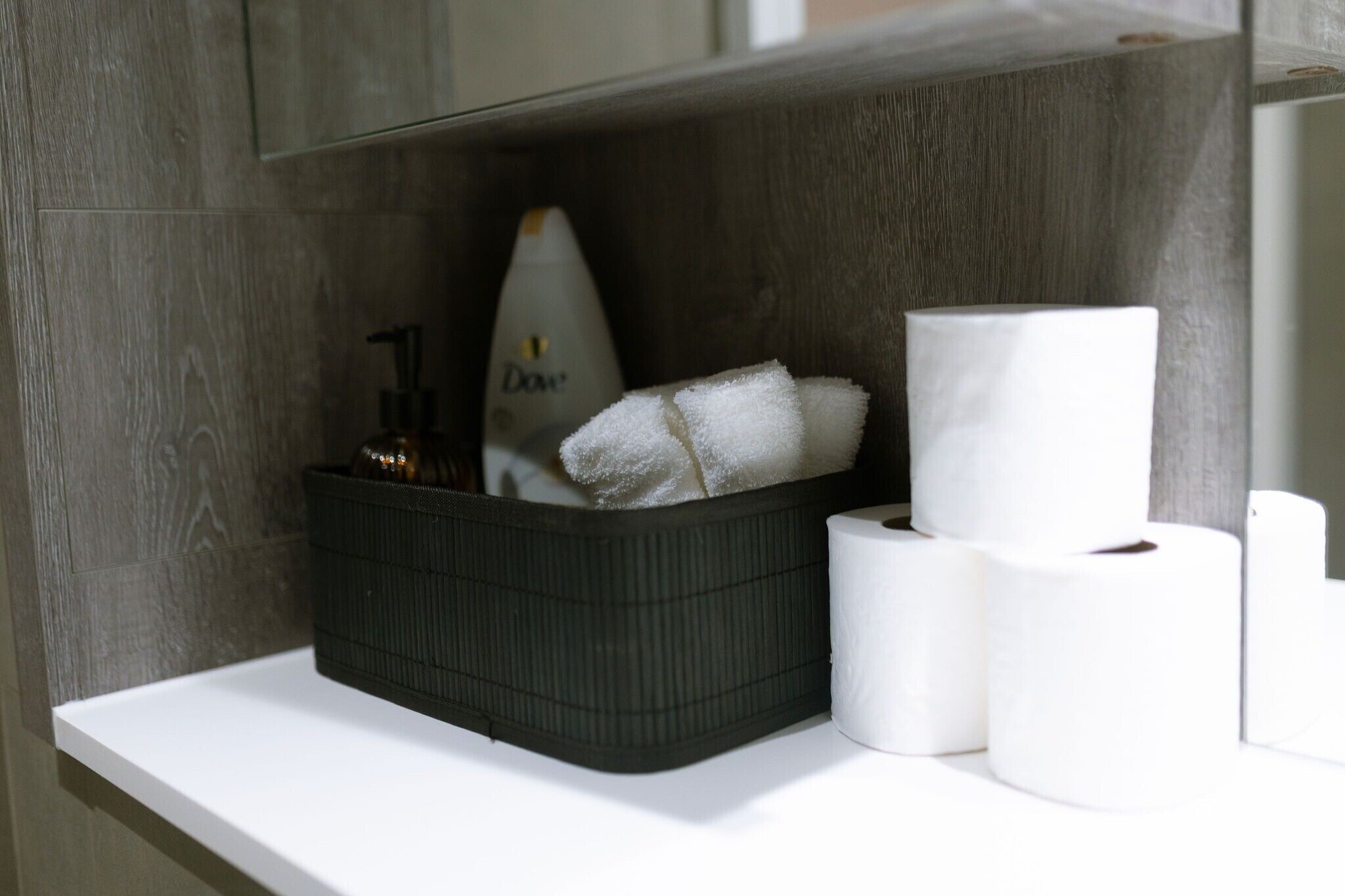 A bathroom shelf with a black basket containing a white towel and a bottle of Dove soap, and two rolls of toilet paper.