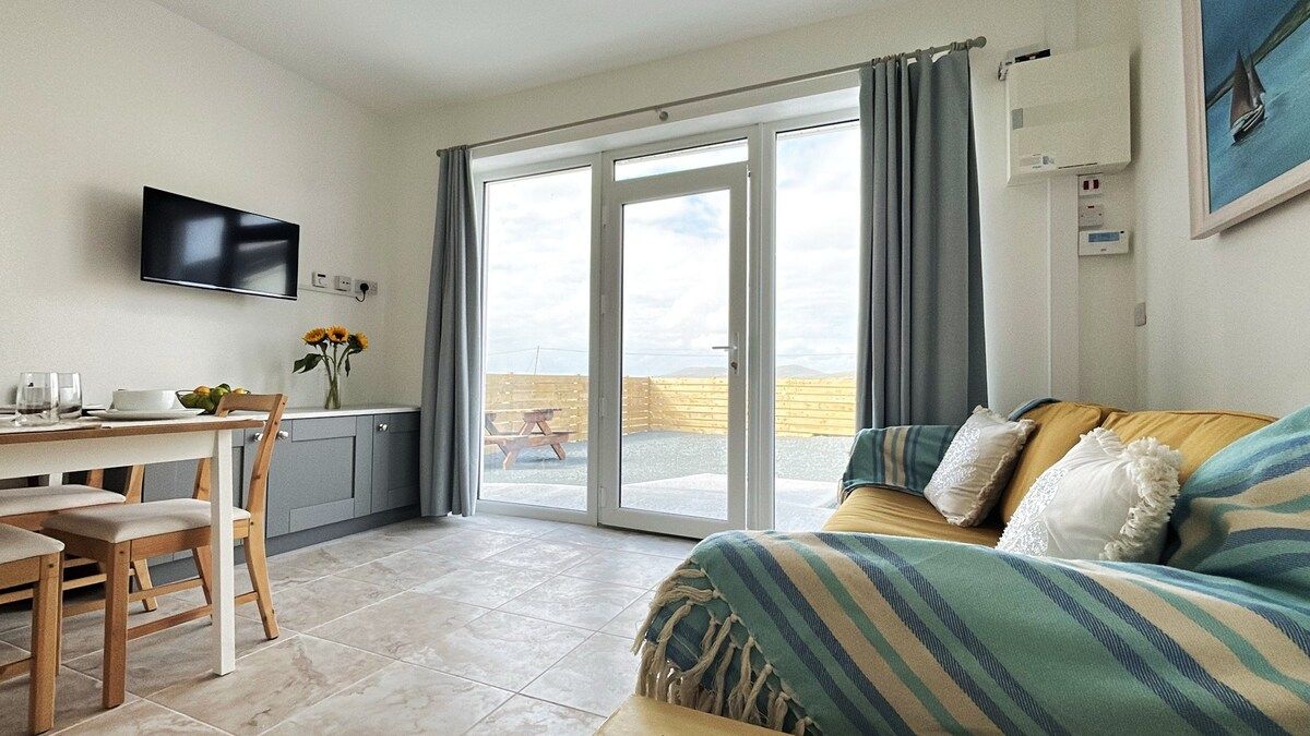 A room with a sofa, a dining table with chairs, a TV mounted on the wall, and a sliding glass door leading to an outdoor area.
