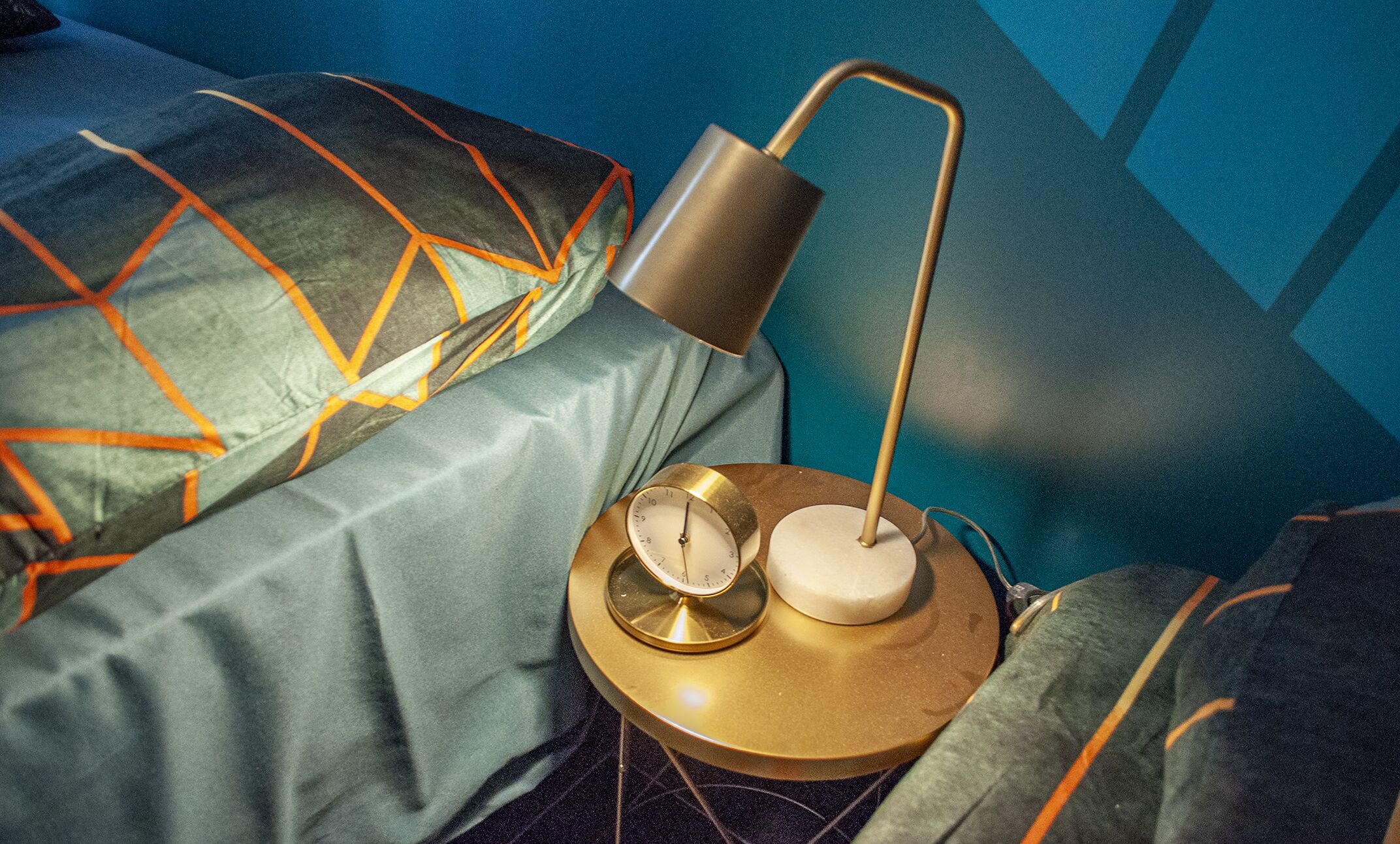 A bed with geometric patterned pillows, a round bedside table with a clock and a lamp, and a blue wall in the background.