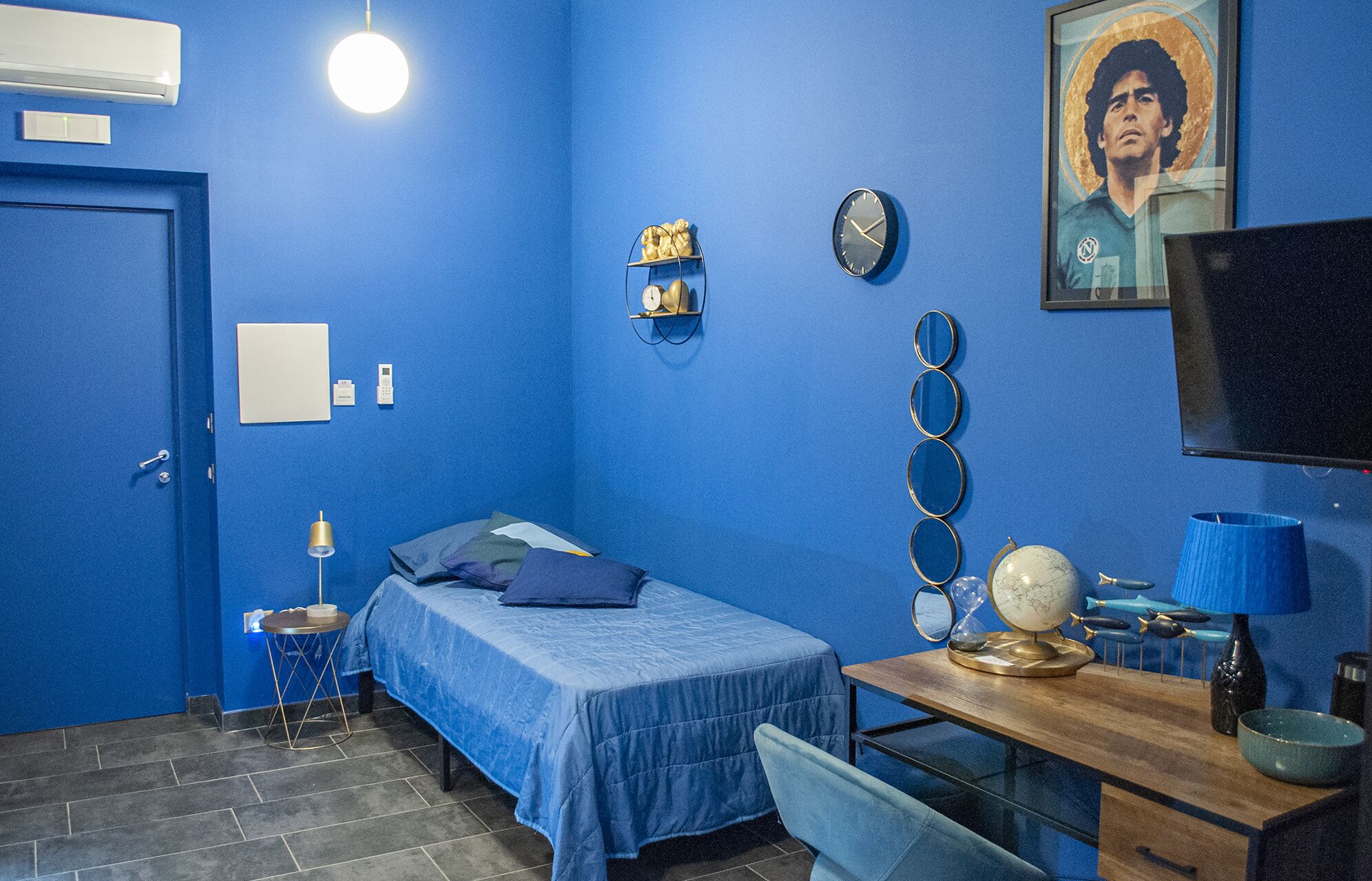 Ein Zimmer mit blauen Wänden, einem blauen Bettbezug, einem Schreibtisch mit Lampe, einer Uhr, einem gerahmten Bild und einem Globus.