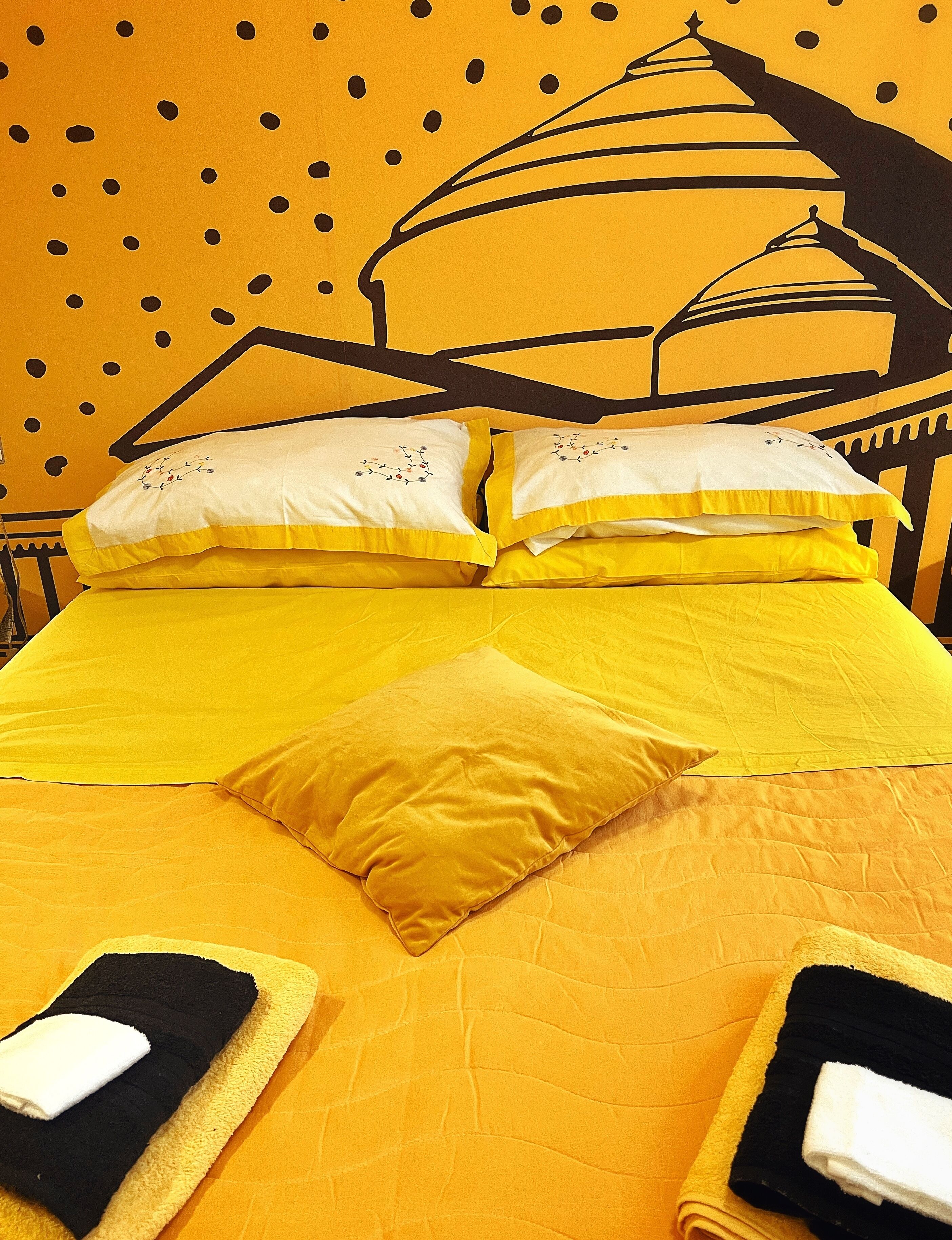 A bed with yellow bedding and pillows, a black and white patterned wall, and a large embroidered design.