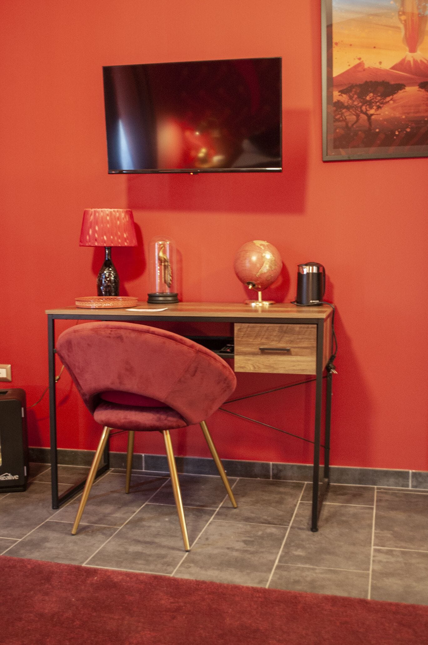 A room with a red wall, a flat-screen TV mounted above a wooden desk, a red velvet chair, a globe, and a coffee maker.