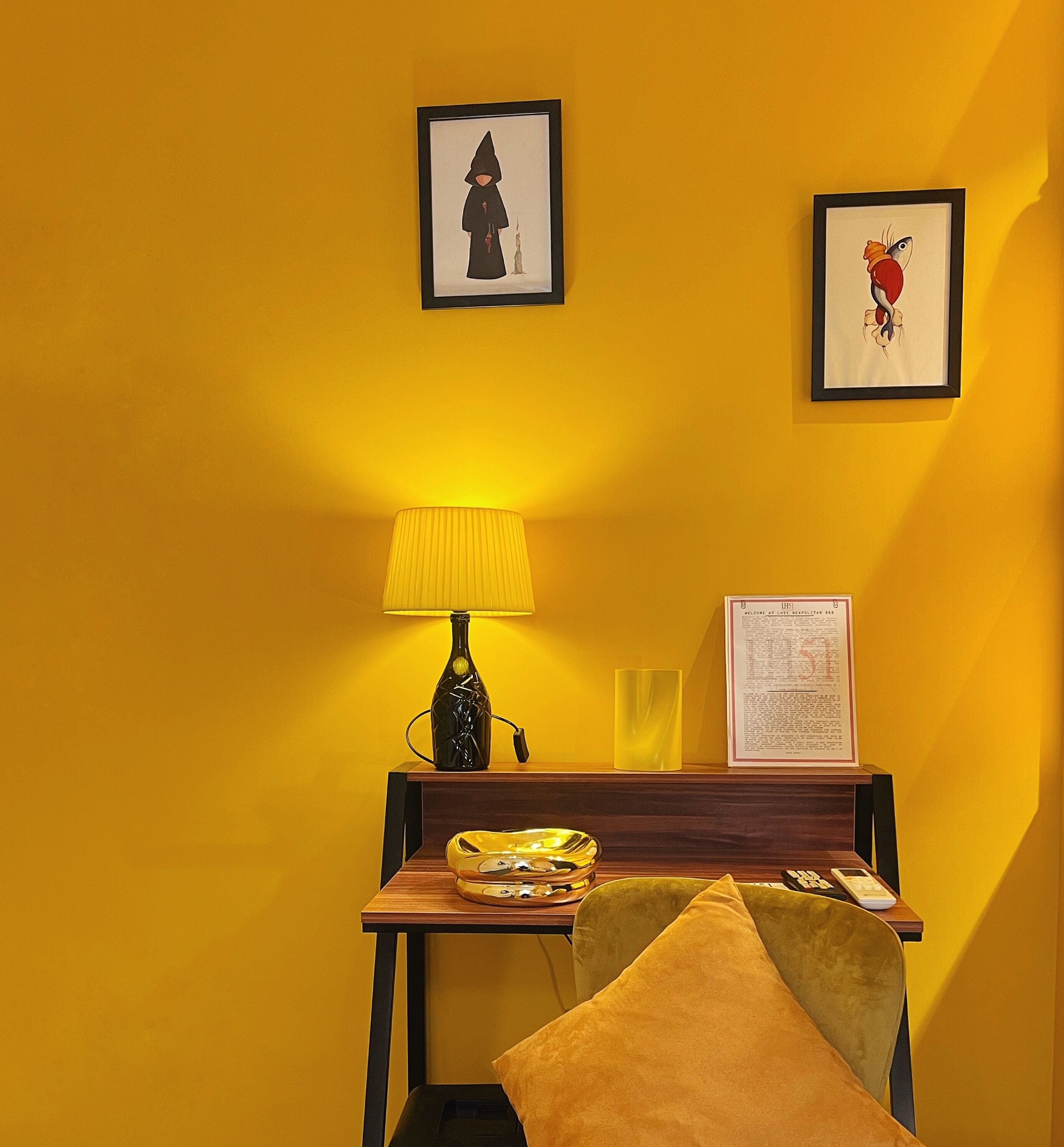 A room with a yellow wall, a wooden table with a lamp, a framed certificate, and a chair with a cushion.