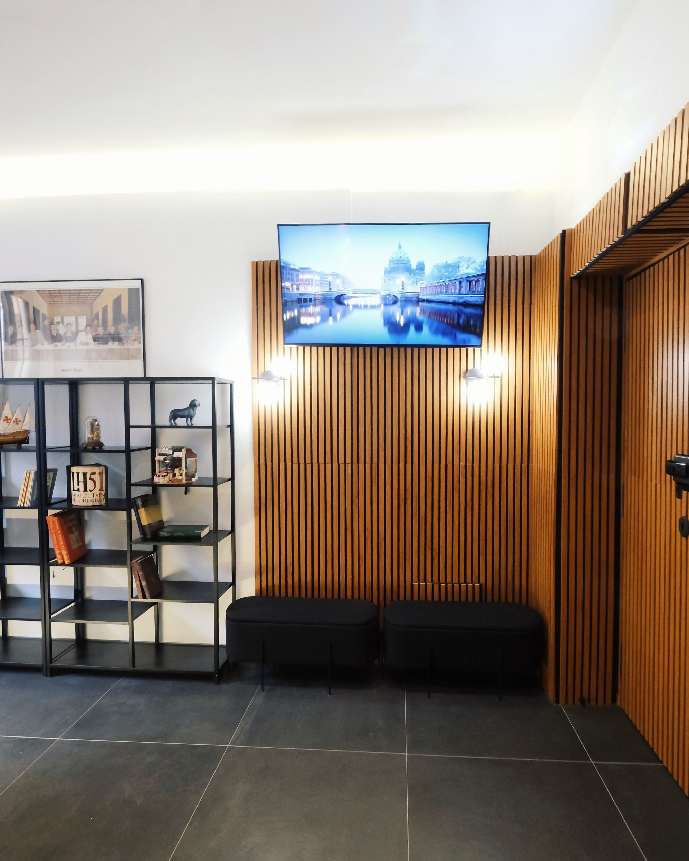 A modern lobby with a large flat-screen TV mounted on a wooden wall, a black shelving unit with decorative items, and a black bench seating area.