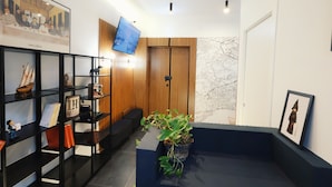 A reception area with a wooden panel wall, a mounted television, a bookshelf with various items, and a potted plant. - LH 51 Neapolitan Rooms (Naples)