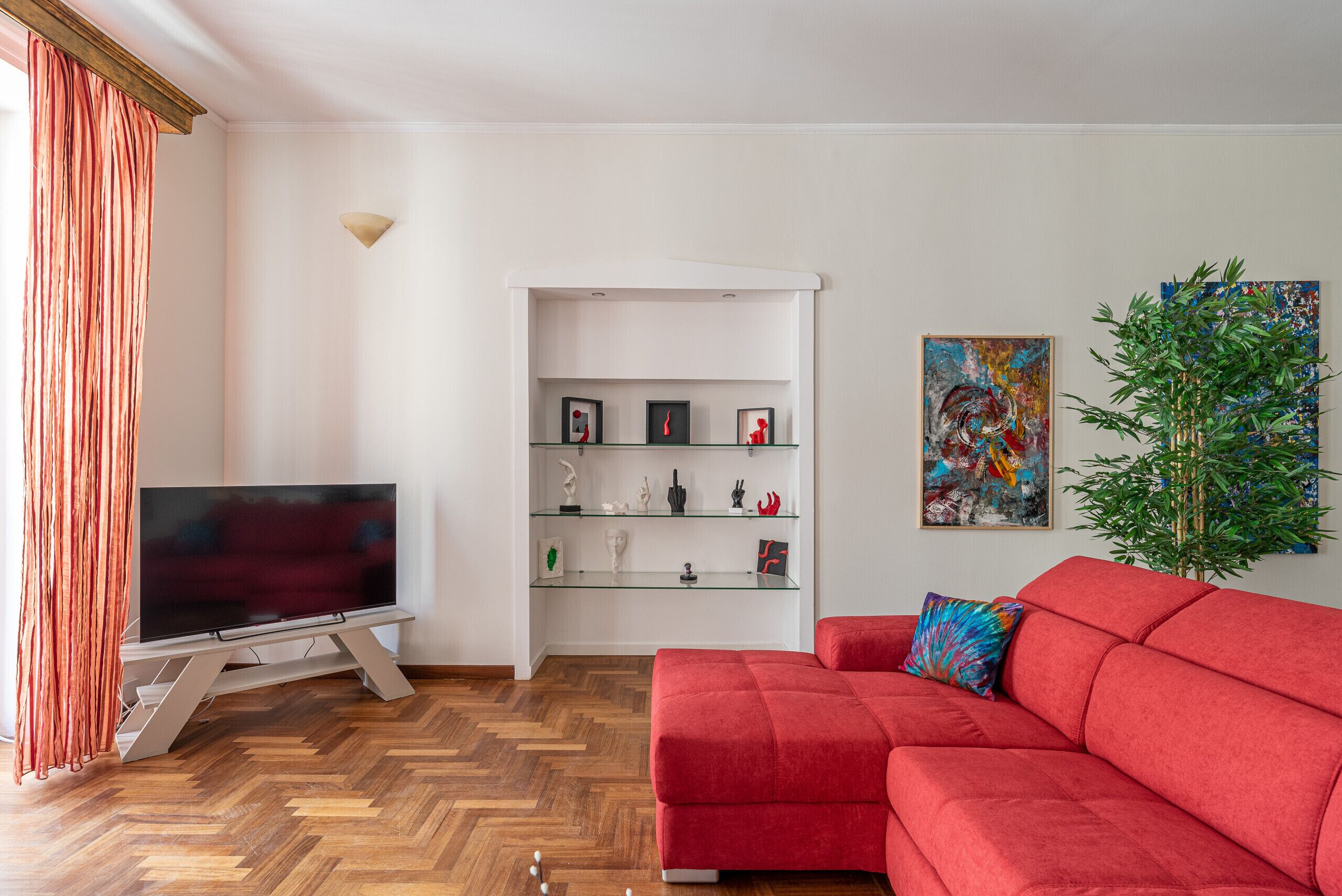 Un salon avec un canapé rouge, un téléviseur posé sur un meuble, une étagère en verre ornée d’objets décoratifs et un tableau accroché au mur.