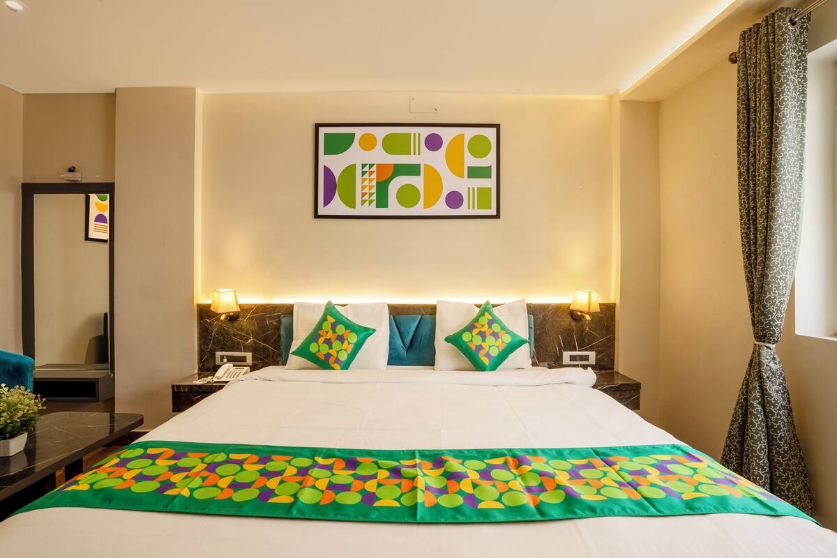 A hotel room with a large bed, two bedside tables with lamps, a mirror, a green and orange patterned bedspread, and a geometric artwork on the wall.