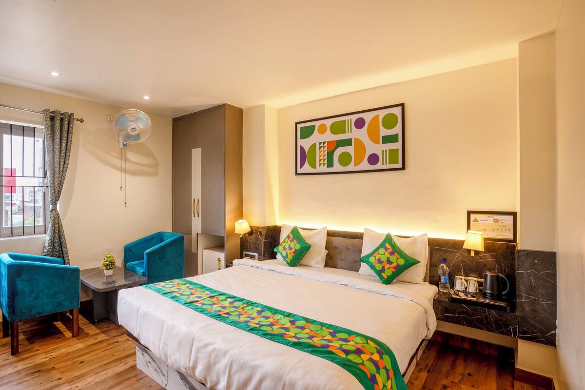 A hotel room with a large bed, a desk with a chair, a standing fan, and a wall-mounted artwork.