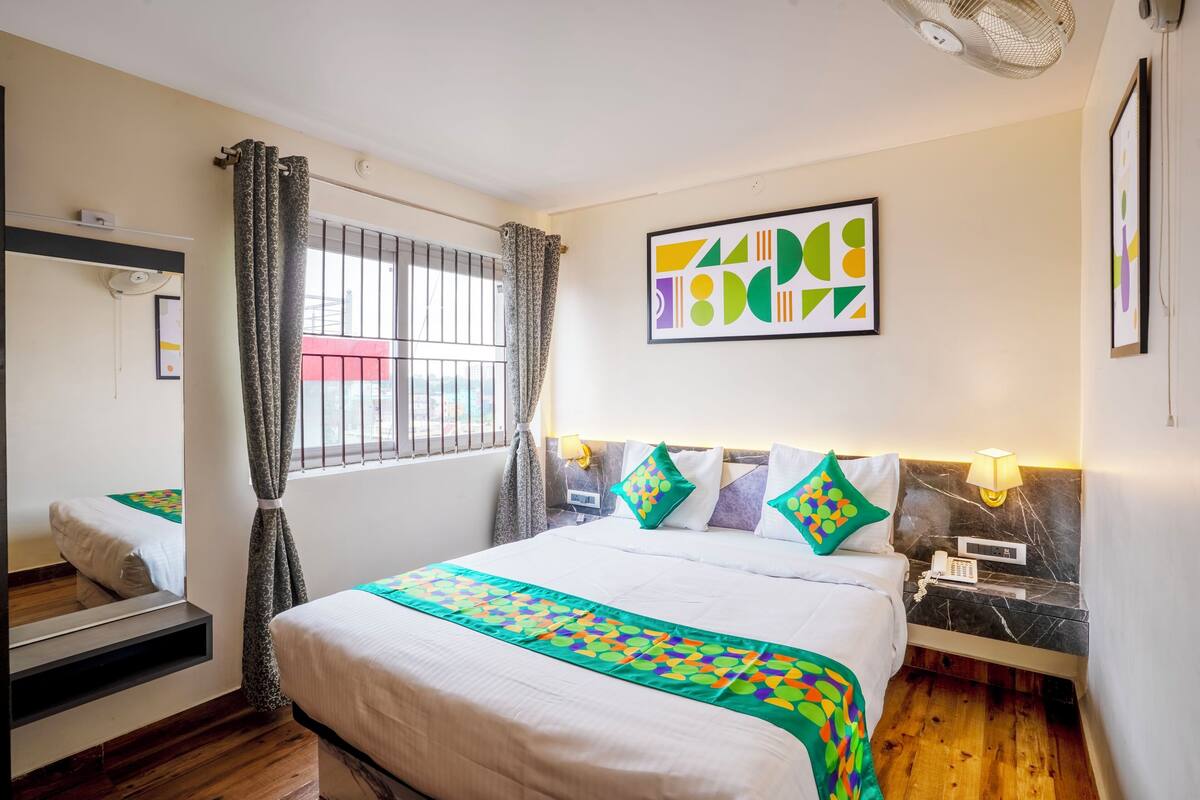 A hotel room with a bed, a bedside table with a lamp, a wall-mounted artwork, and a window with curtains.