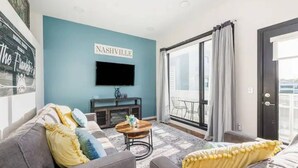 A living room with a blue accent wall, a flat-screen TV mounted above a wooden console table, a sofa with yellow and blue cushions, and a large sliding glass door leading to an outdoor area. - Experience the best of Nashville from this stunning downtown condo, just 1.2 miles from Broadway’s live music, restaurants, and nightlife. Featuring a resort-style pool, and secure garage parking. (Nashville)