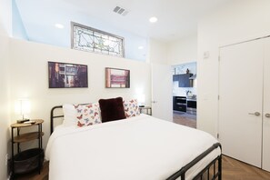 A bedroom with a bed, a bedside table with a lamp, a small shelf, and a wall-mounted TV. There is a door leading to a kitchen area.