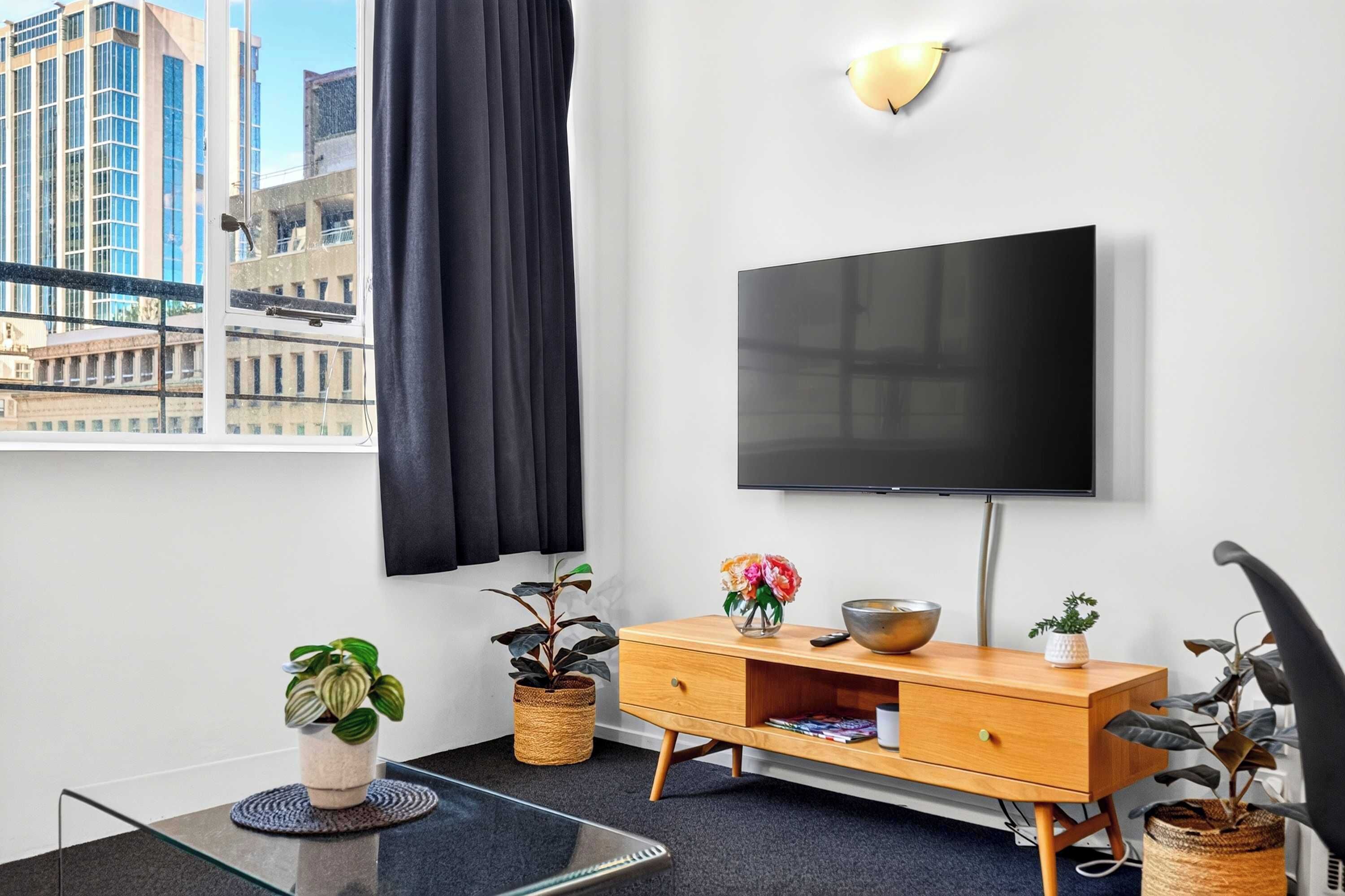 Ett modernt vardagsrum med en stor platt-TV som är monterad på väggen, en träskommod, ett soffbord i glas och utsikt över stadsbilden genom ett fönster.