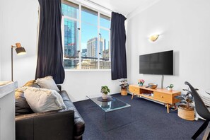 Ein modernes Wohnzimmer mit einer Couch, einem Couchtisch, einem Fernseher und Blick auf die Stadt durch große Fenster.