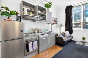 A compact kitchen with a refrigerator, oven, microwave, and built-in dishwasher. There is a sofa and a small table with a plant. - Stay on Iconic Queen St by Zodiak Stays (Auckland)