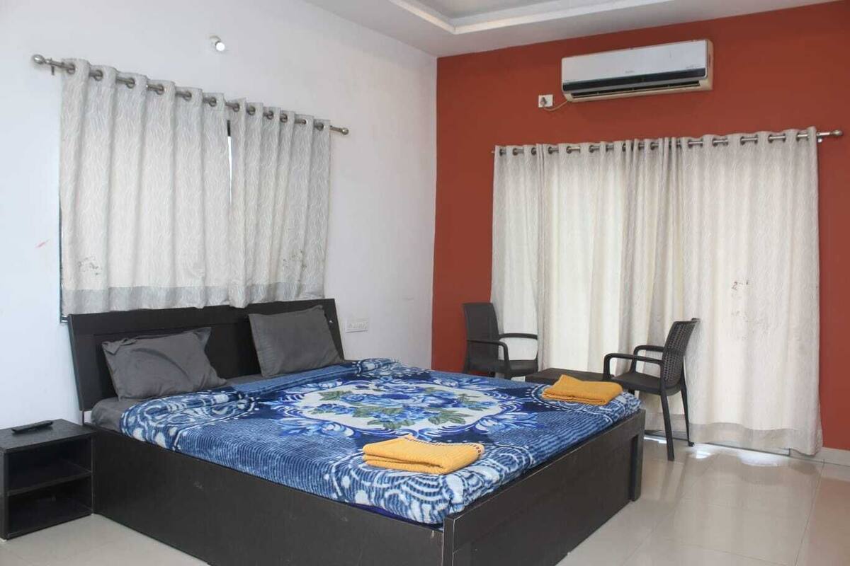 A bedroom with a bed, two chairs, an air conditioner, and curtains.