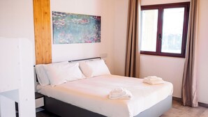 A neatly made bed with white linens and a folded towel, a wooden headboard, a painting on the wall, and a window with curtains.