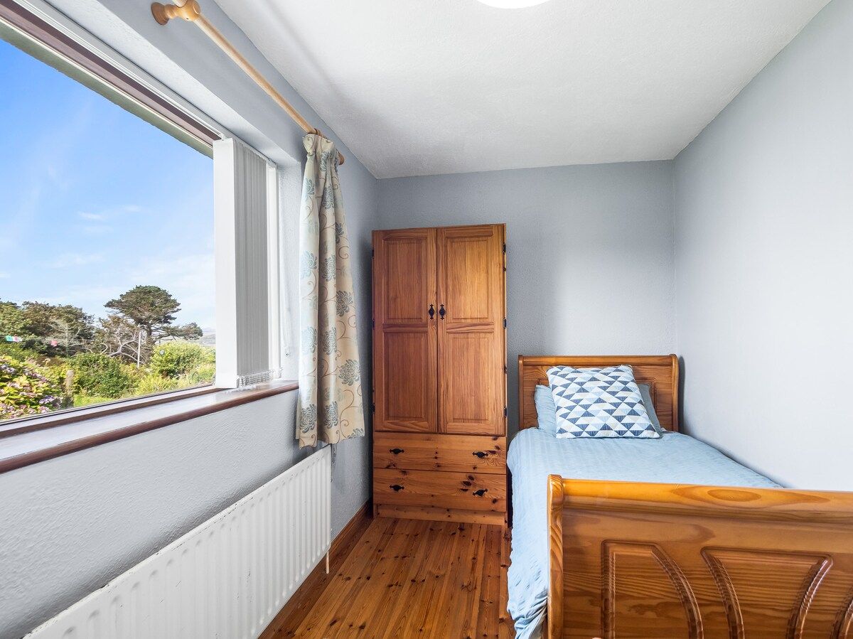Ein Schlafzimmer mit einem Holzschrank, einem Bett mit einem gemusterten Kissen und einem Fenster mit Blick auf Grünflächen.