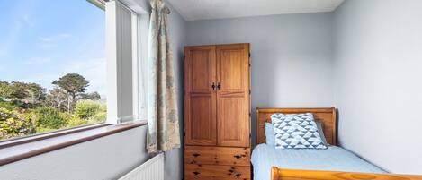 Ein Schlafzimmer mit einem Holzschrank, einem Bett mit einem gemusterten Kissen und einem Fenster mit Blick auf Grünflächen.
