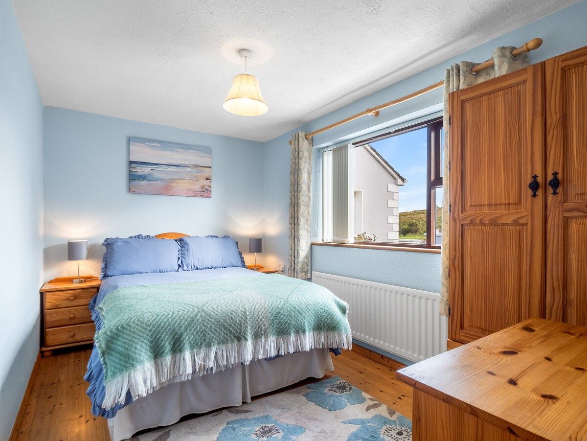A bedroom with a bed, bedside lamps, a window with curtains, a wooden table, and a painting on the wall.