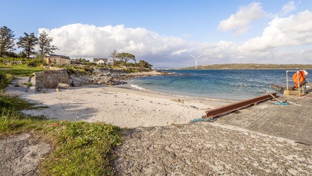 A beach with a rocky shoreline, a wind turbine, and a few houses in the background.