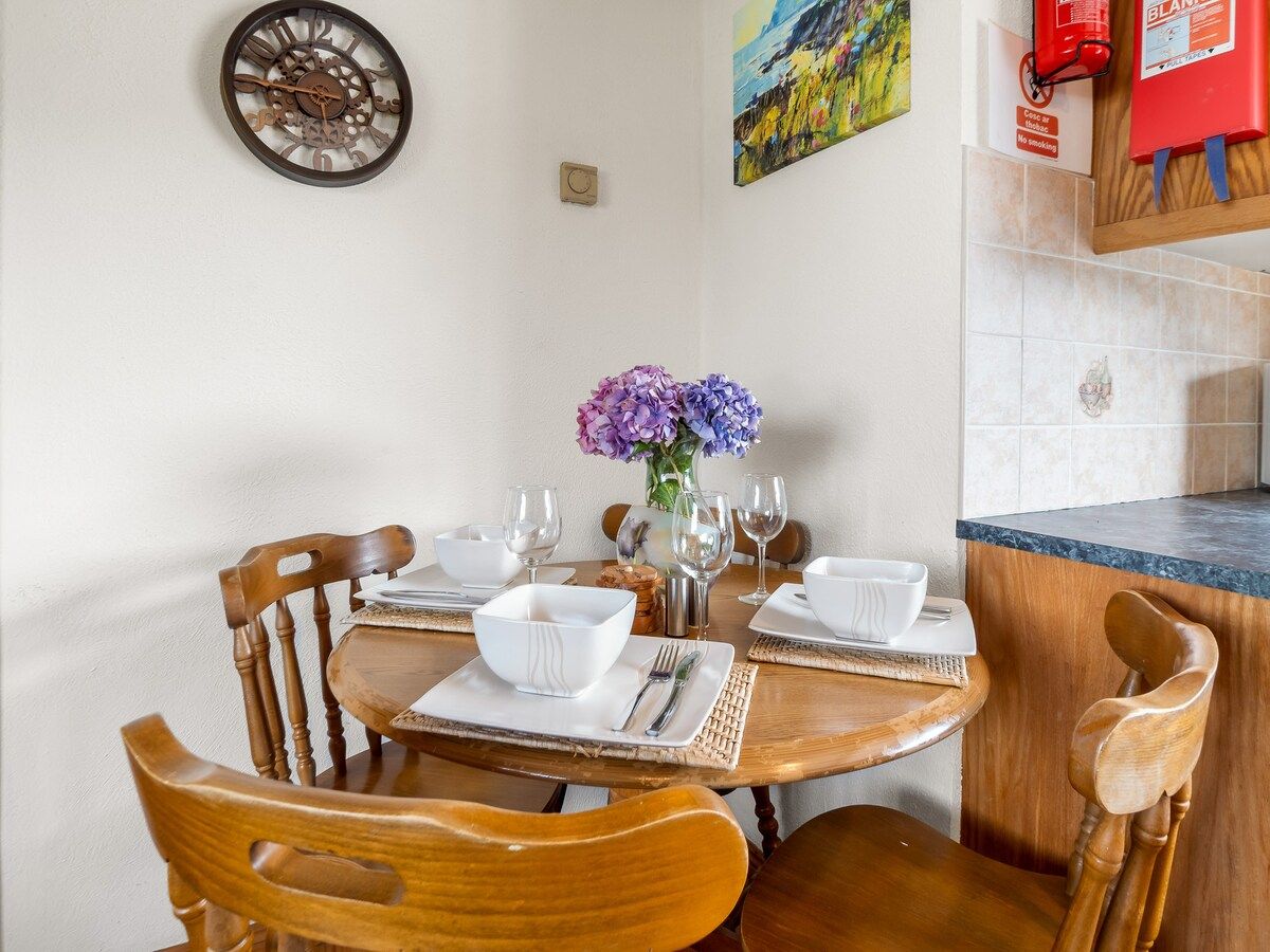 Un coin repas avec une table en bois dressée pour deux personnes, un vase de fleurs et une horloge murale.