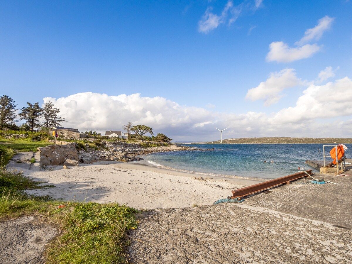 A coastal beach with a clear sky, a sandy shore, and a wind turbine in the distance.