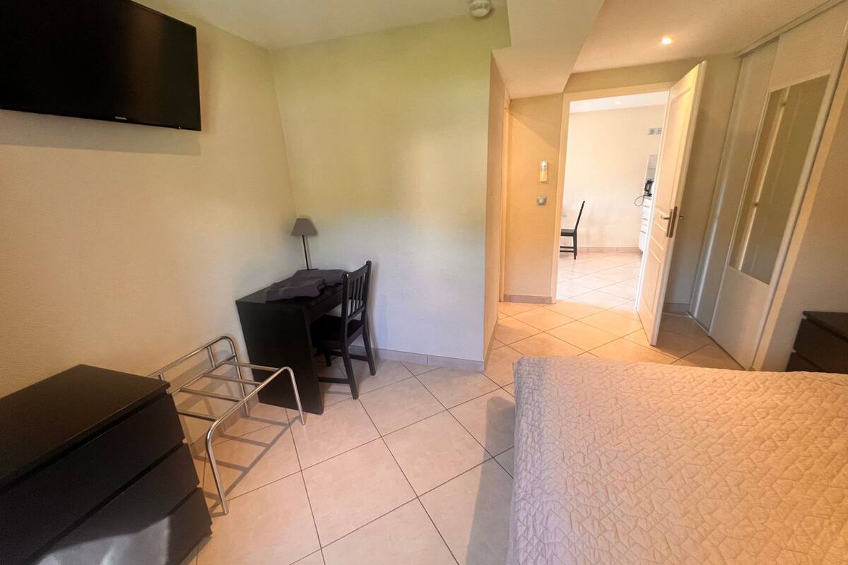 A bedroom with a bed, a desk, and a chair. There is a TV mounted on the wall and a door leading to another room.